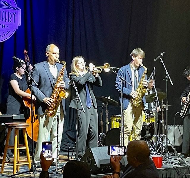 Live jazz band on stage with saxophones, trumpet, drums, and a microphone under blue-purple lights