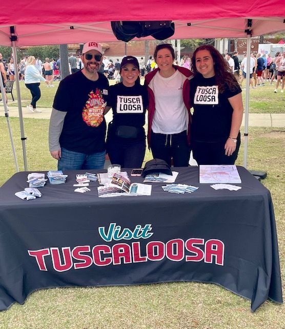 Four people at a Tuscaloosa booth under a pink canopy, with a “Visit Tuscaloosa” table banner.