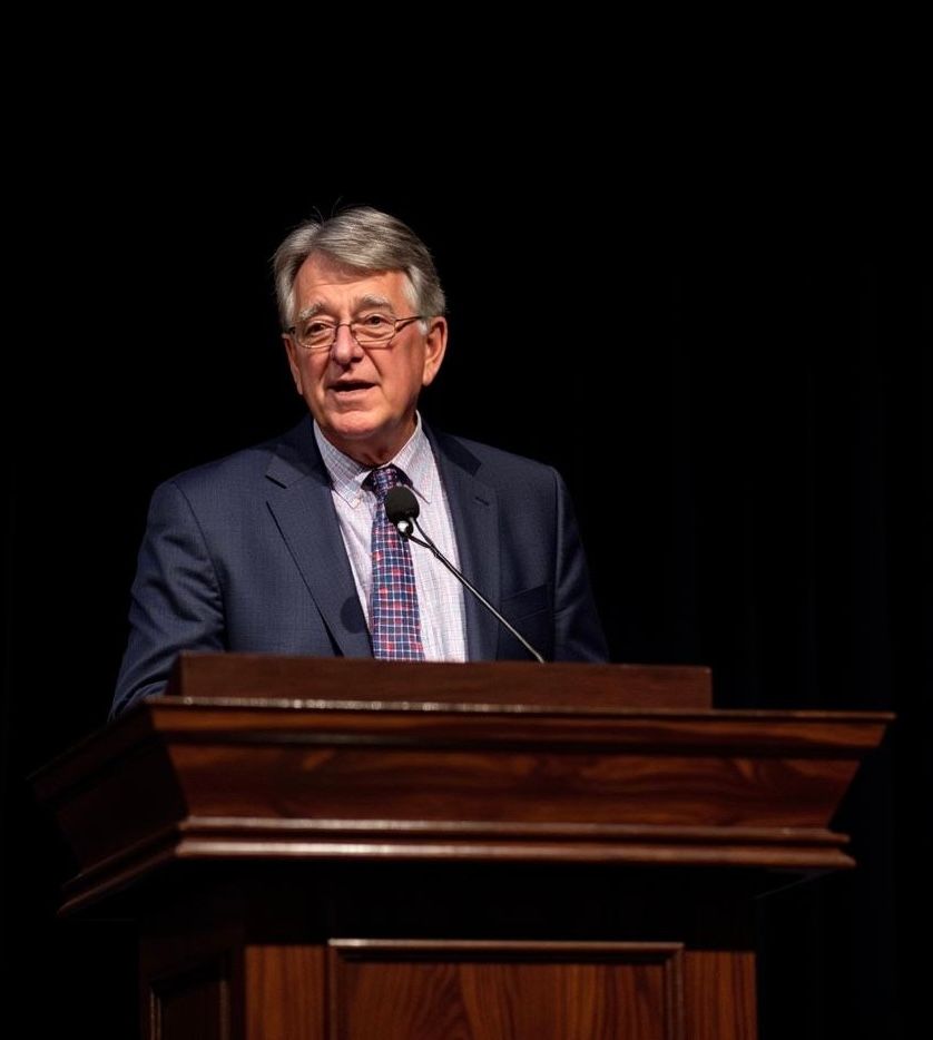 Man speaking at a podium on a dark stage, wearing a suit and tie.