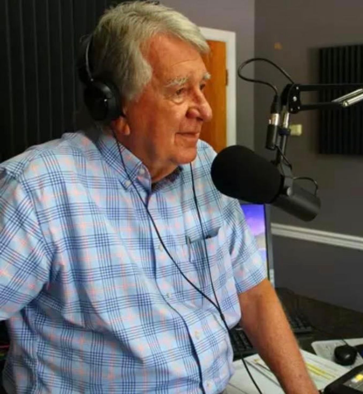 Man wearing headphones speaking into a microphone in a radio studio, seated beside a desk