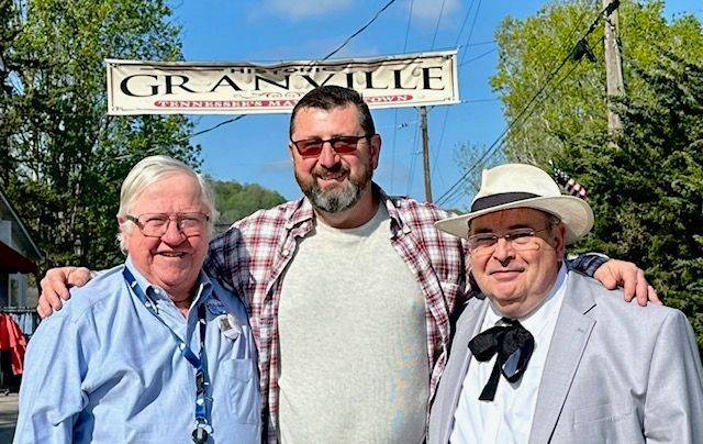 Three men smiling outdoors under a “Granville” sign, one with arms around the others.