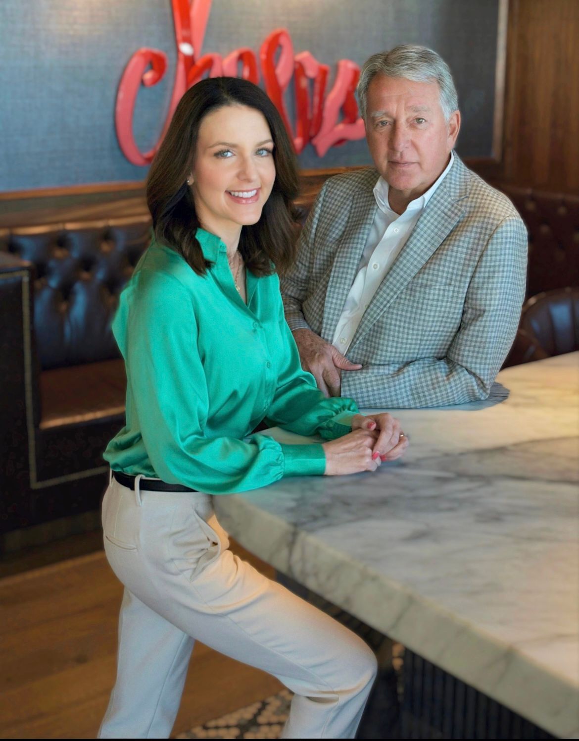Two people posing at a marble bar counter in a warmly lit restaurant lounge with a red neon sign behind them