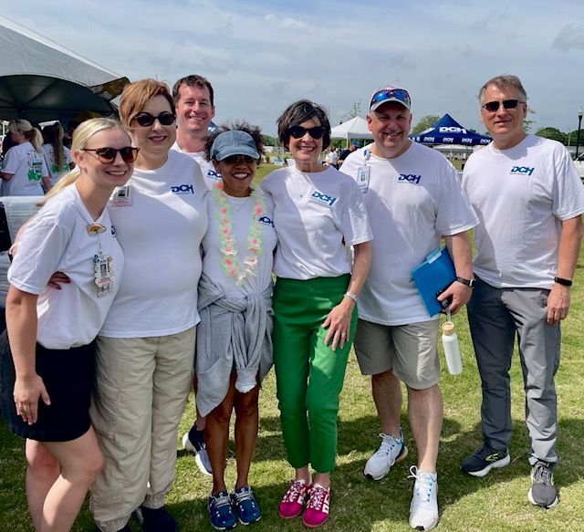 Seven people in white shirts stand together outdoors on a sunny grassy field at an event