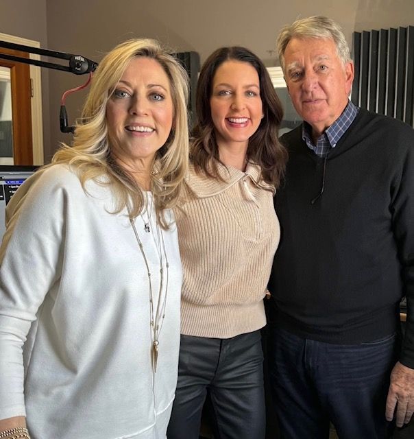Three people smiling indoors beside a microphone in a studio setting.