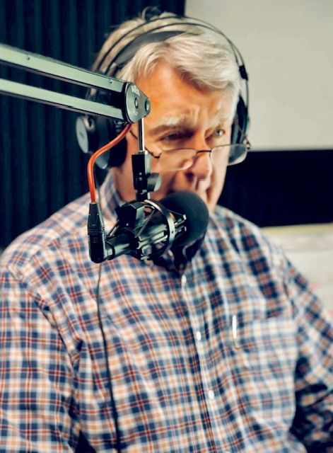 Man wearing headphones speaks into a studio microphone, with a plaid shirt and focused expression.