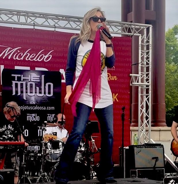 Singer onstage at an outdoor concert, wearing sunglasses and a pink scarf, singing into a microphone.