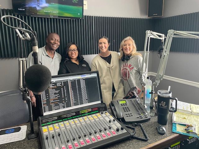 Four people smiling in a radio studio beside a mixing console and microphone.