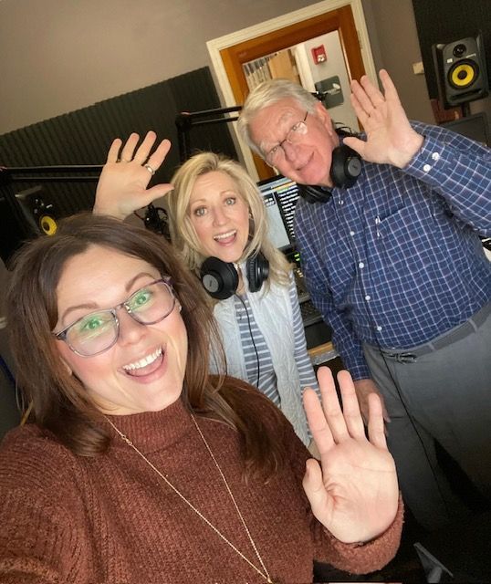 Three people waving in a recording studio, smiling at the camera.