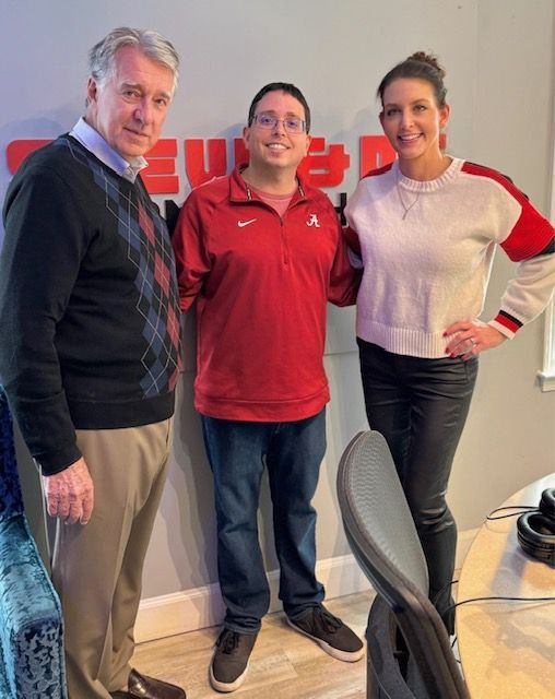 Three people posing in a radio studio, standing beside a chair and microphone.