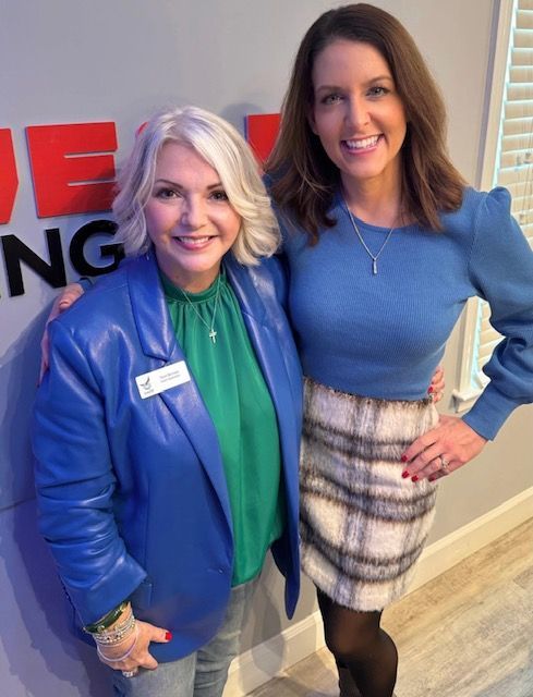 Two women smiling indoors, one in a blue blazer and green blouse, the other in a blue top and plaid skirt.