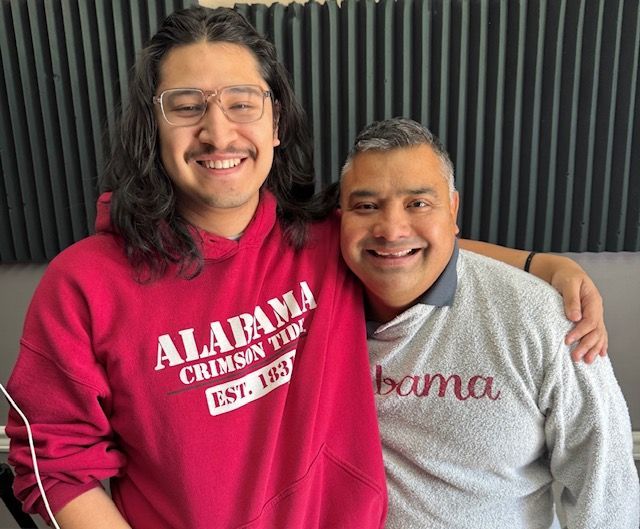Two smiling people in Alabama sweatshirts posing indoors against a dark curtain backdrop.