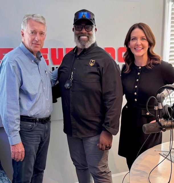 Three people stand in a radio studio beside microphones and a control table, smiling at the camera.