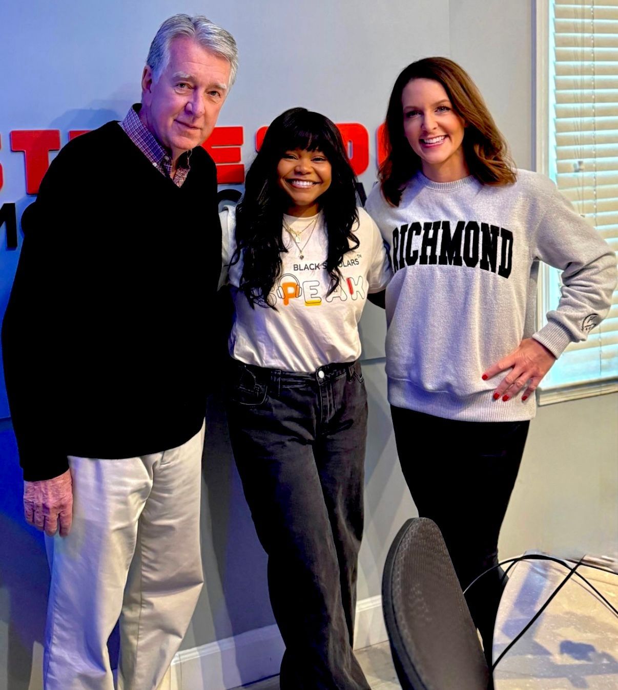 Three people posing indoors, two in sweatshirts, with a window and chairs in the background.