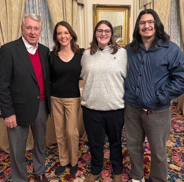 Four people standing indoors, smiling in front of gold curtains and a framed mirror.