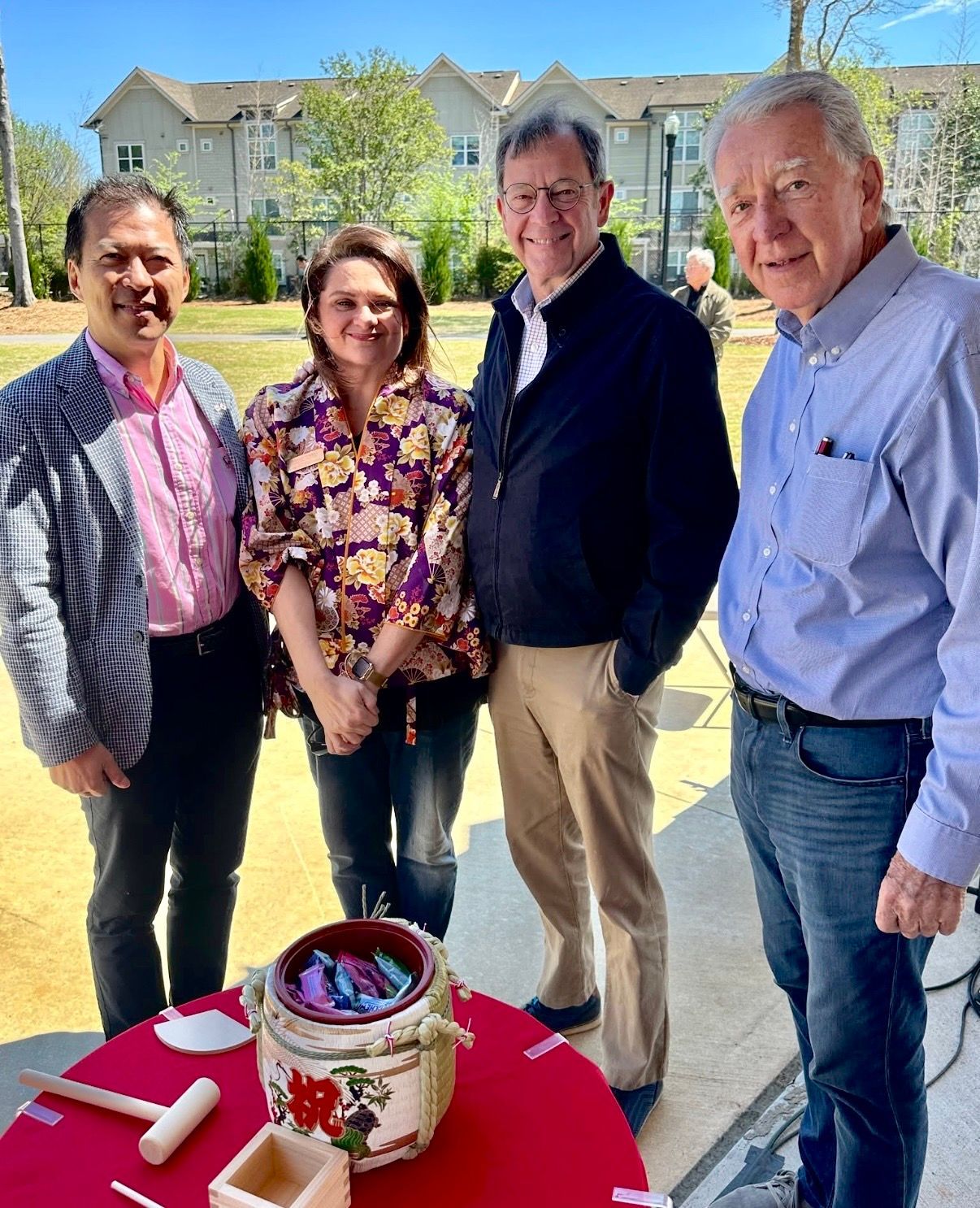 Four people stand outdoors beside a red table with a decorated bucket, smiling in front of houses.