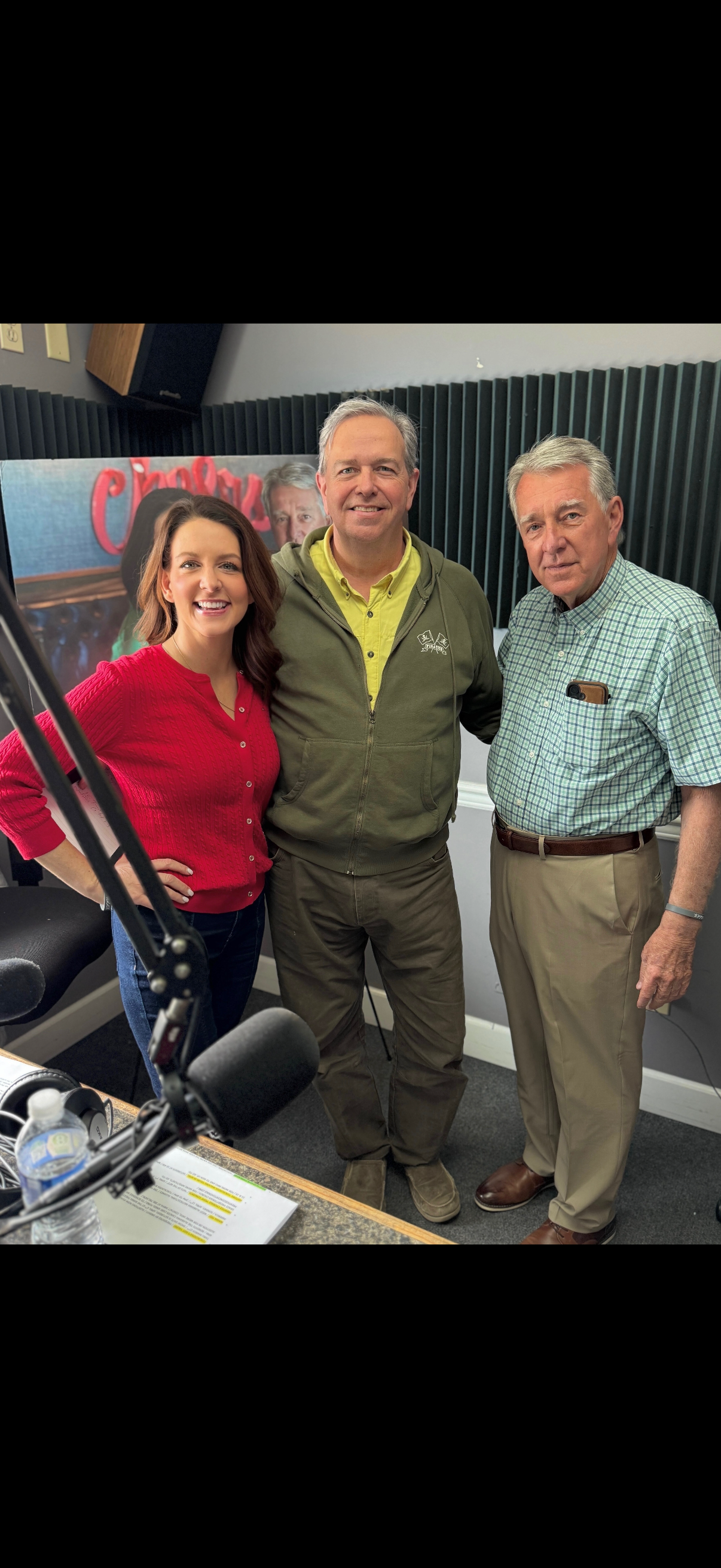 Three people smiling in a radio studio, standing beside a microphone and audio equipment.
