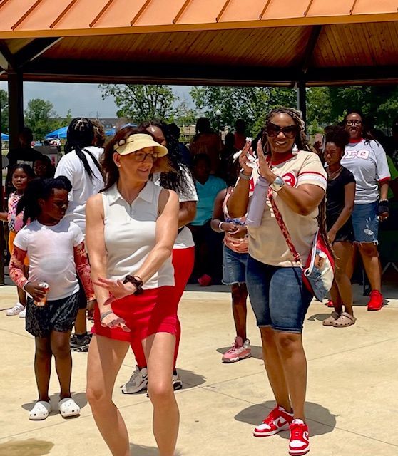 People walking outdoors under a pavilion, with a woman in red shorts and visor in the foreground.
