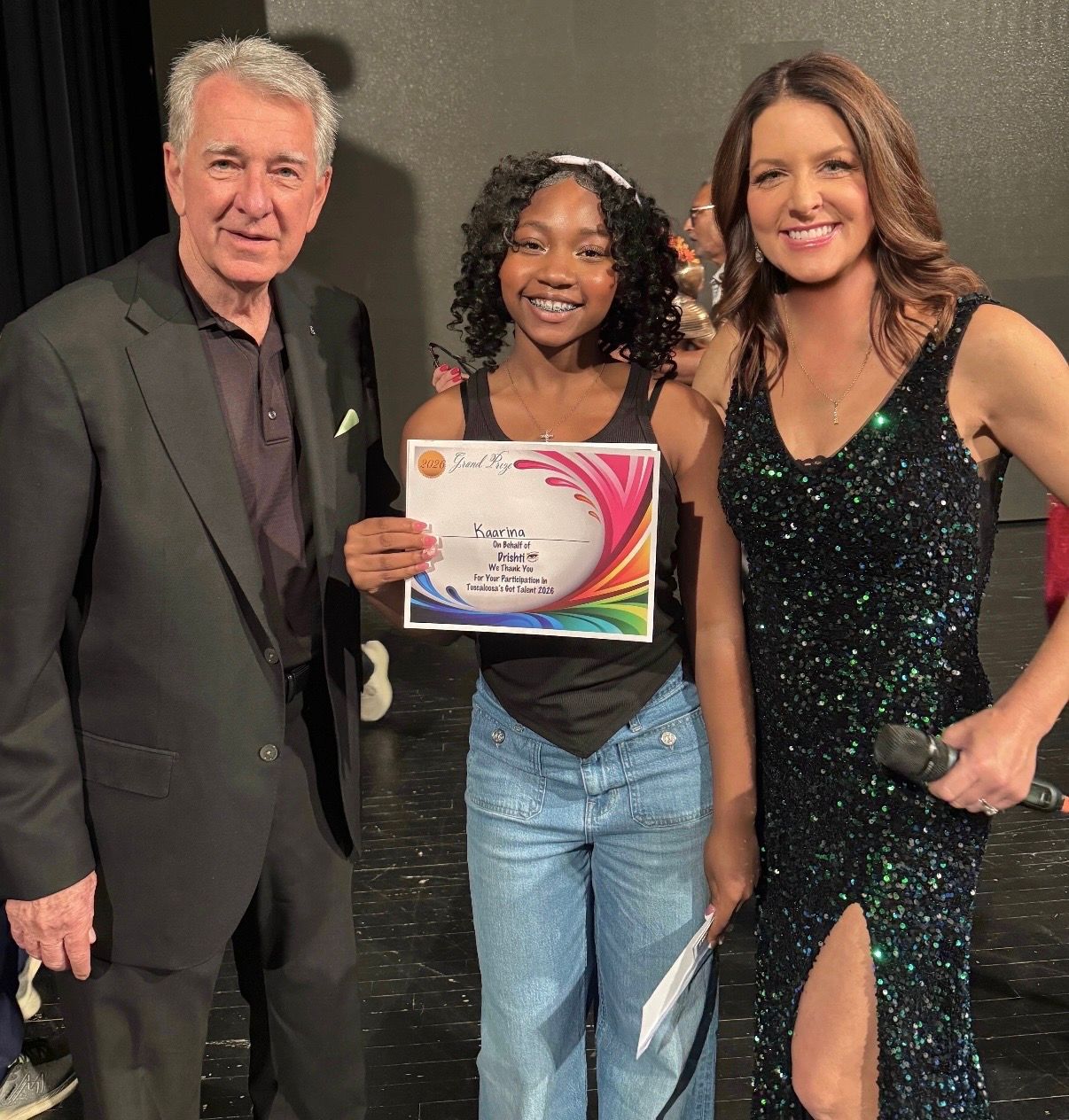 Three people posing at an event, one holding a certificate, two in black formal attire.