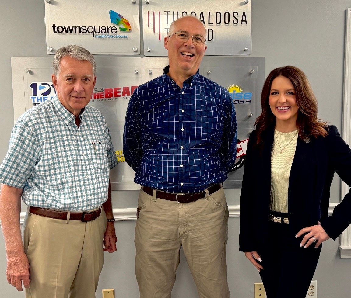Three people posing indoors in front of a wall with signs and logos.