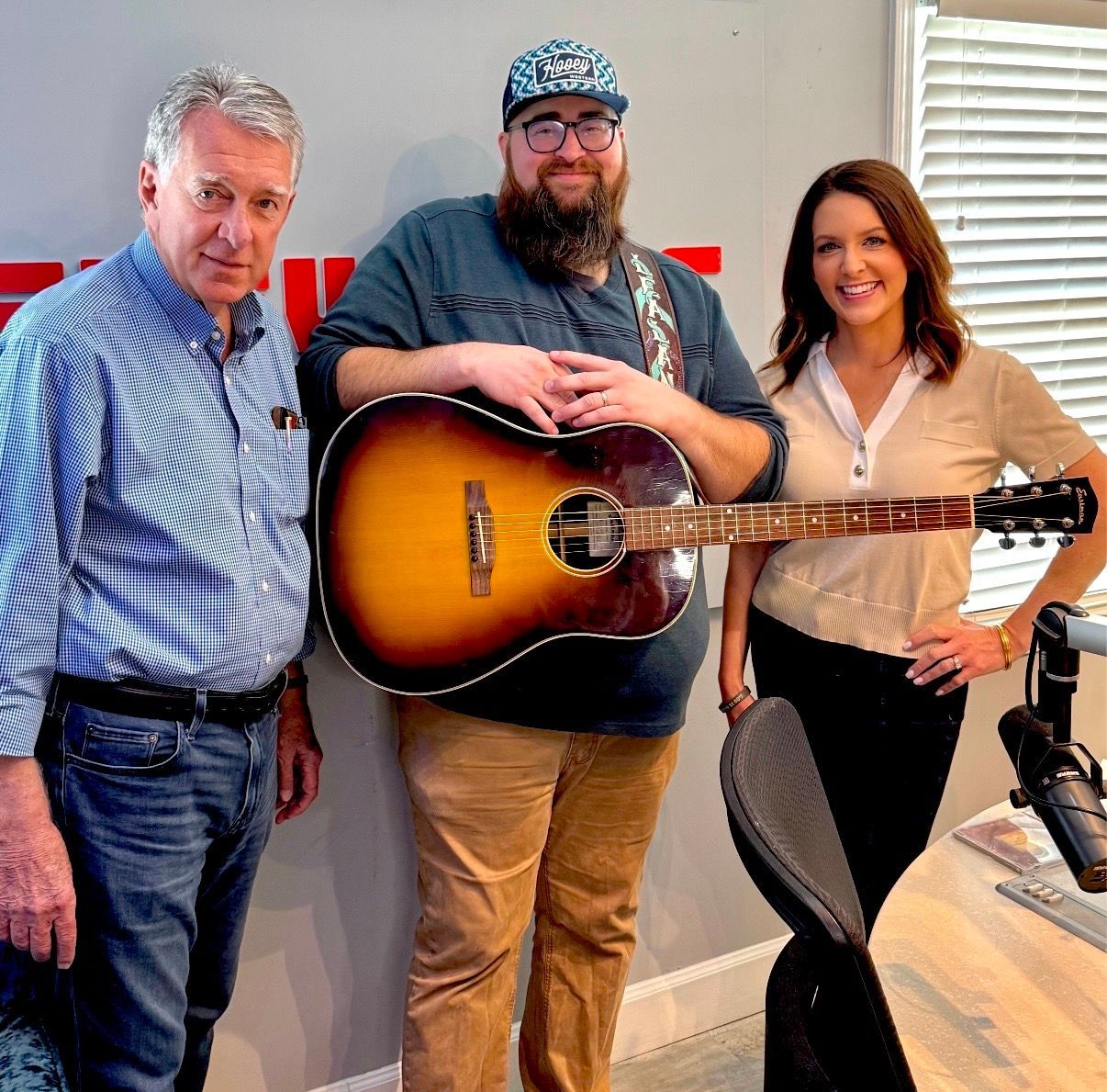 Three people standing in a studio, one holding a guitar, smiling at the camera.