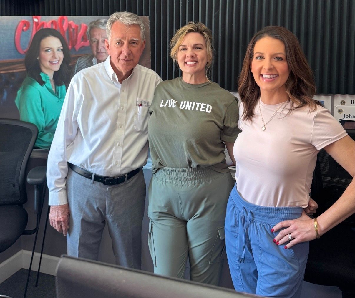 Four people posing indoors, two in “United” shirts, in front of a wall photo.