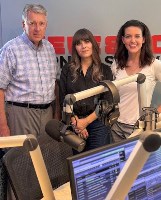 Three people standing in a radio studio with microphones and mixing equipment, smiling at the camera.