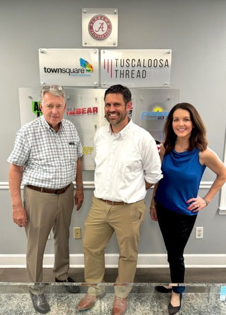 Three people posing indoors in front of company logos, smiling at the camera.