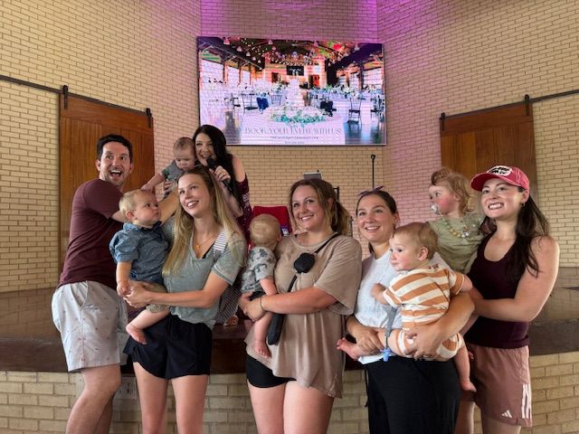 Group of people posing indoors in front of a wall-mounted screen, smiling and holding two children.