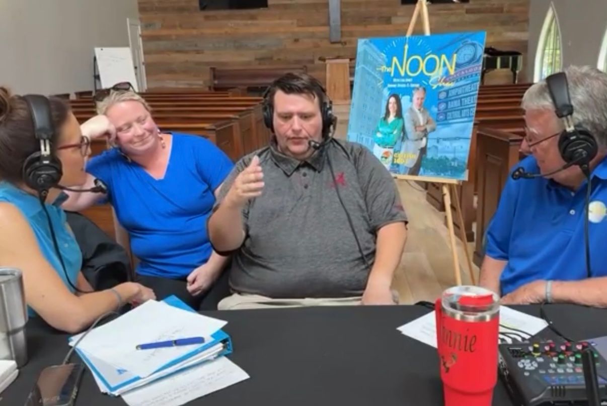 Four people wearing headphones at a podcast table with microphones and papers, one speaking into a mic