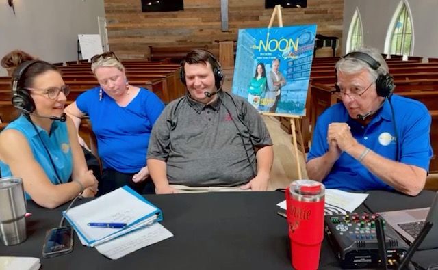 Four people wearing headsets listen around a table with papers, a laptop, and a red tumbler in a church hall