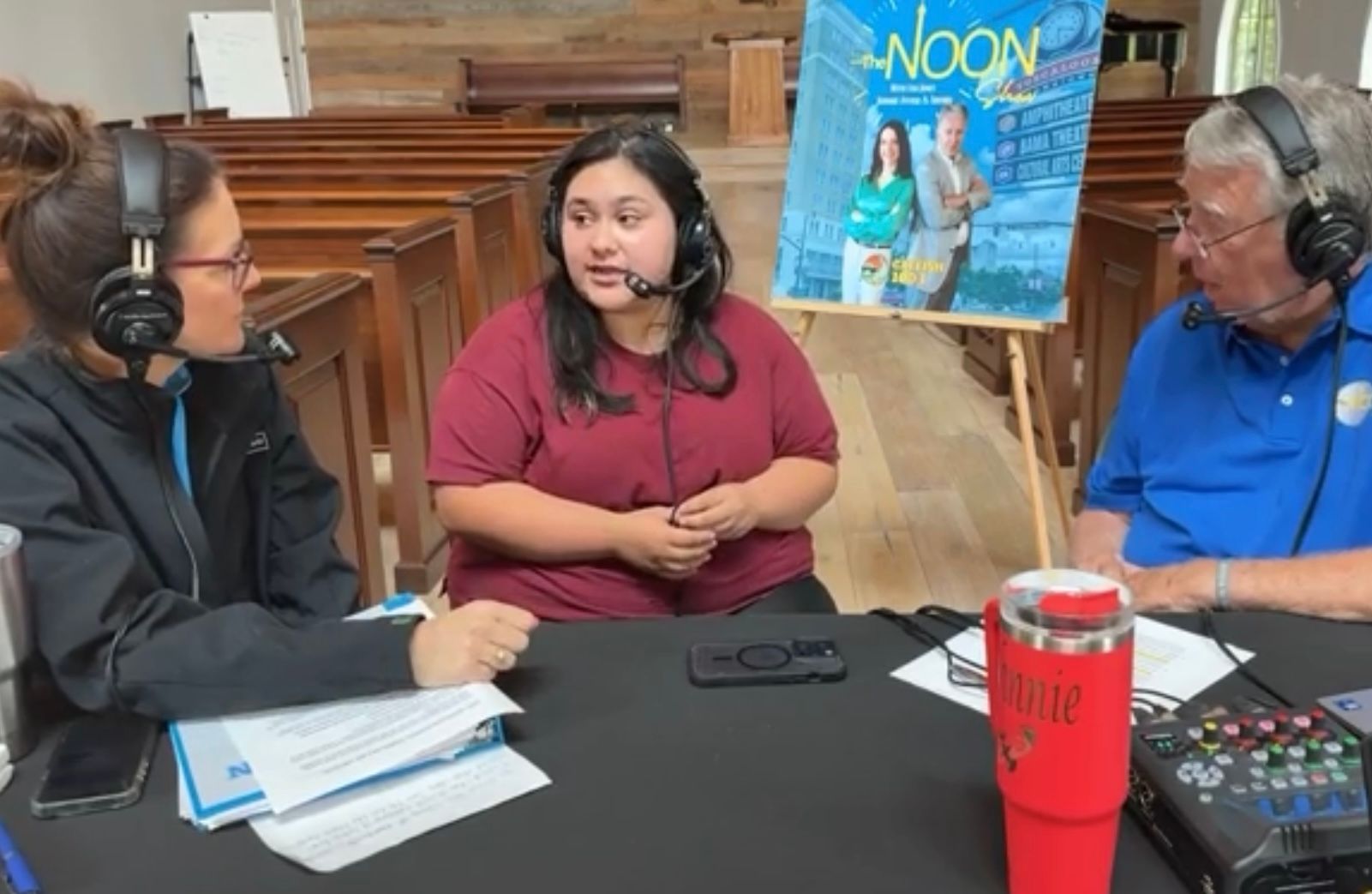 Three people wearing headsets record a podcast at a table with microphones and notes.