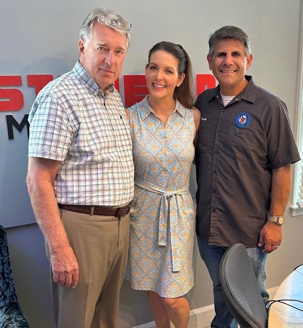 Three smiling people standing together indoors beside a wall with a logo.