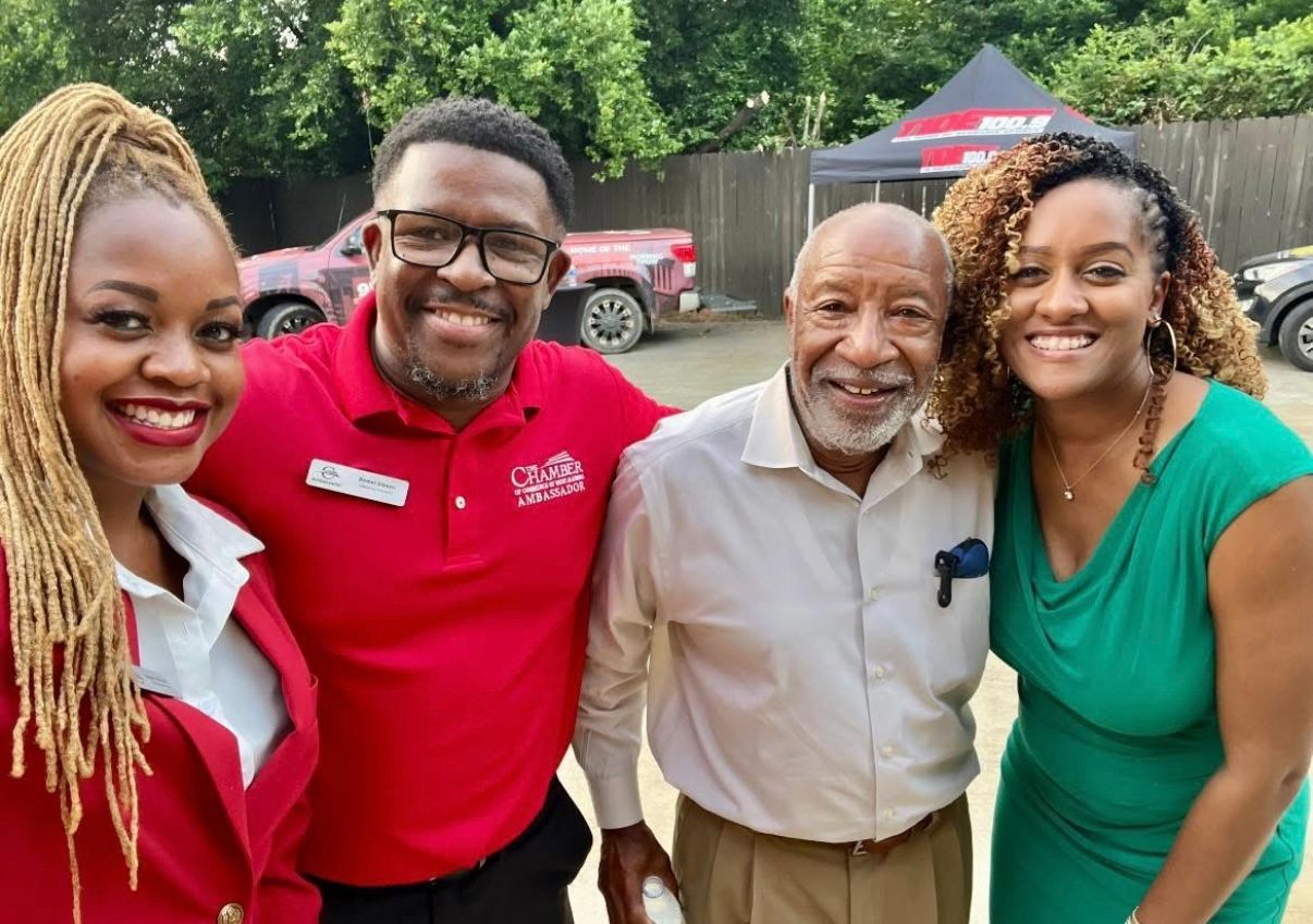 Four smiling people posing outdoors at a park event, with tents and trees in the background.
