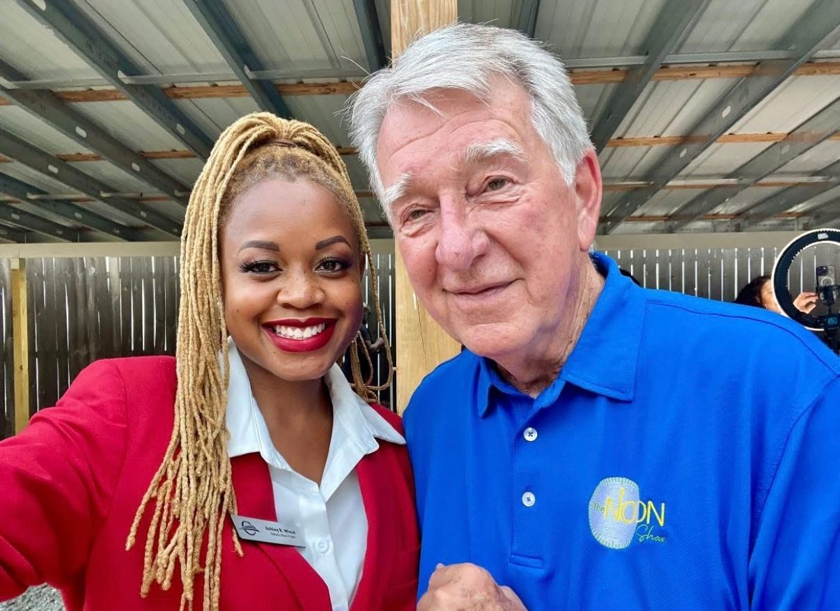 Two smiling people posing outdoors, one in a red jacket and one in a blue polo shirt.