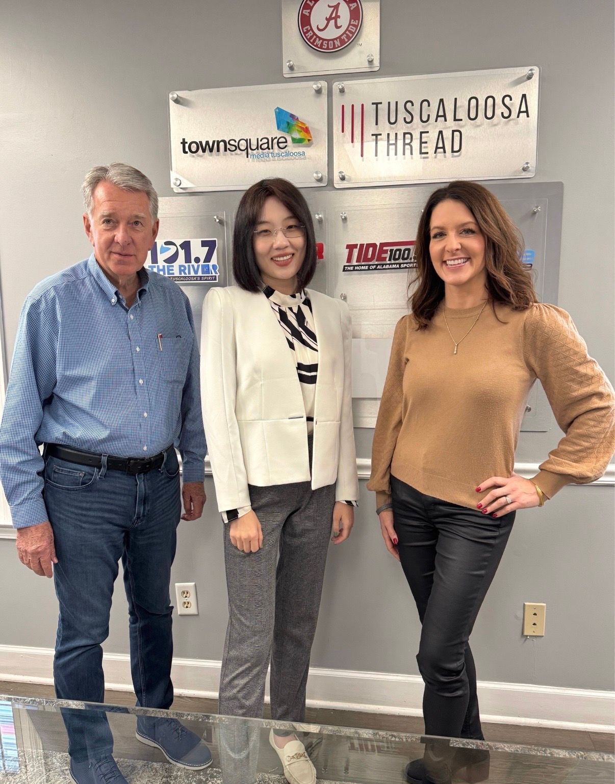 Three people posing indoors in front of Tuscaloosa Thread signs, smiling at the camera.