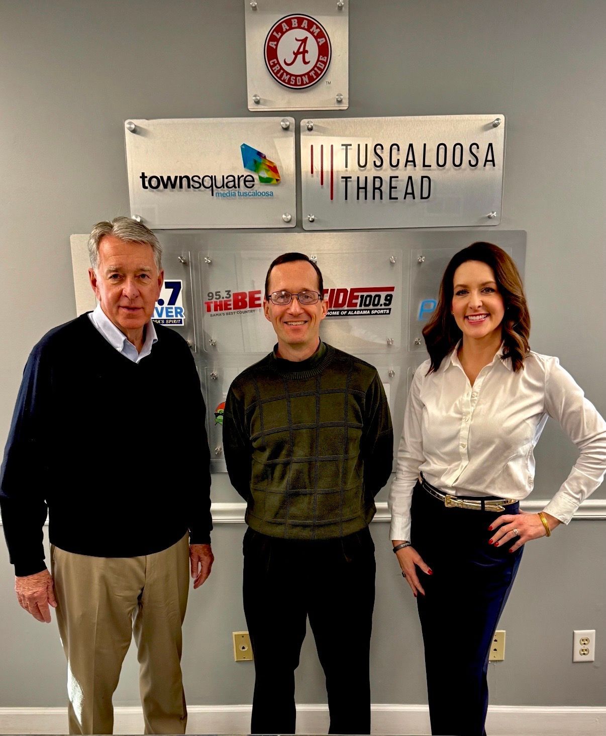 Three people posing indoors in front of company logos, smiling at the camera.