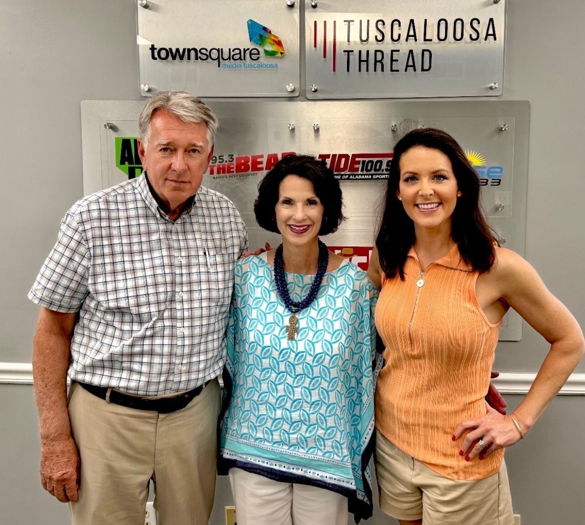 Three people posing indoors before a Tuscaloosa Thread sign, smiling at the camera.