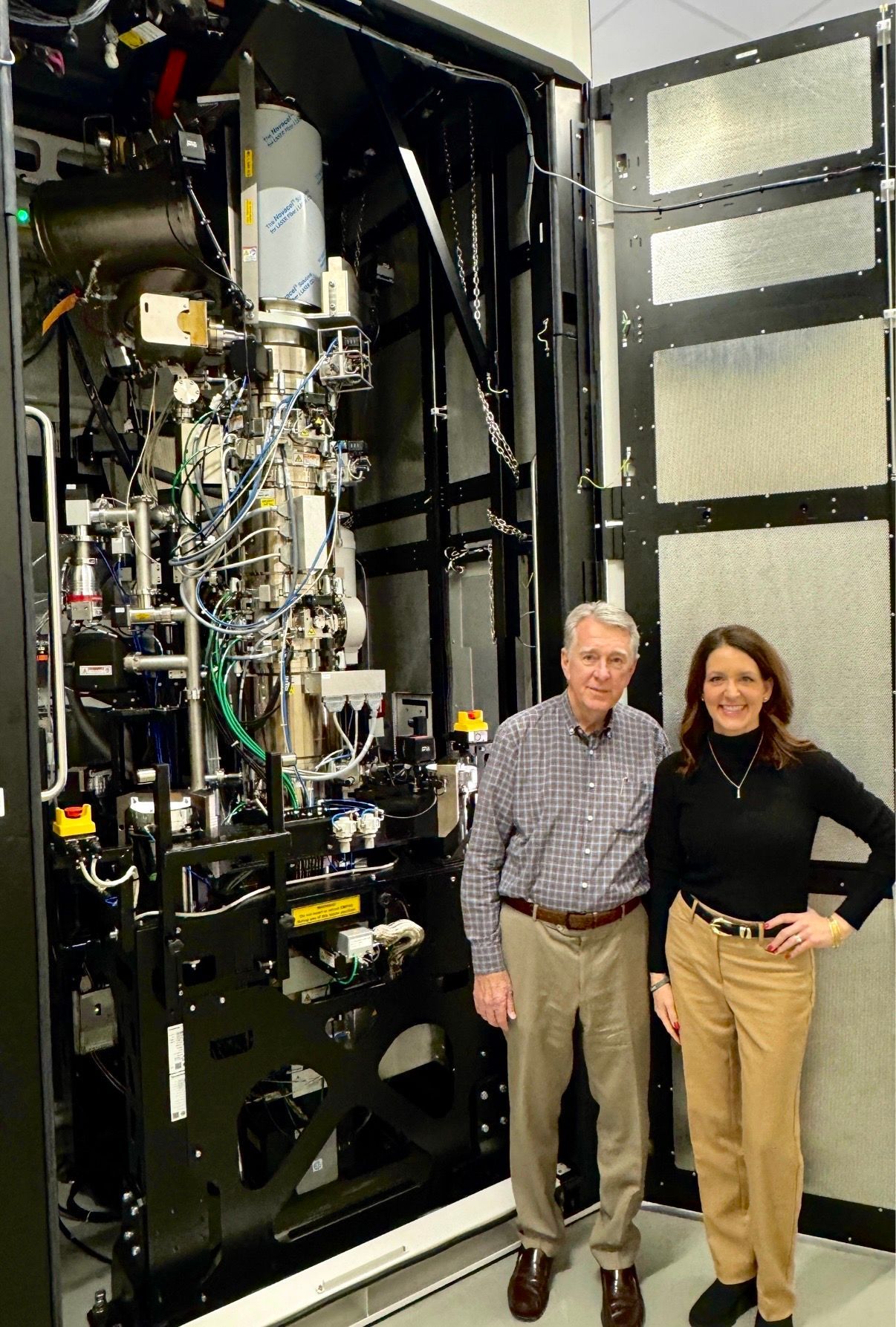 Two people standing beside a large black scientific machine in a lab-like room