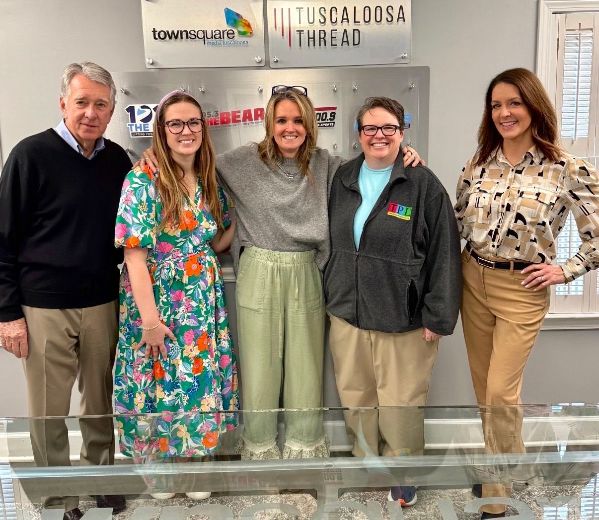 Five people posing indoors in front of a Tuscaloosa Thread sign, smiling at the camera.