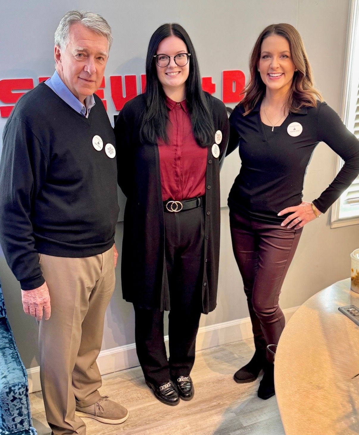 Three people pose indoors, smiling, in front of a wall with red lettering.