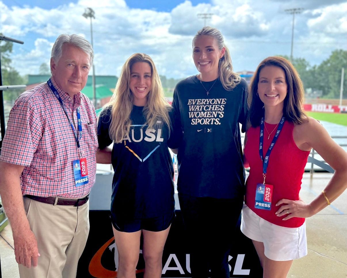 Four people posing outdoors at a sports event, wearing lanyards and casual team shirts.