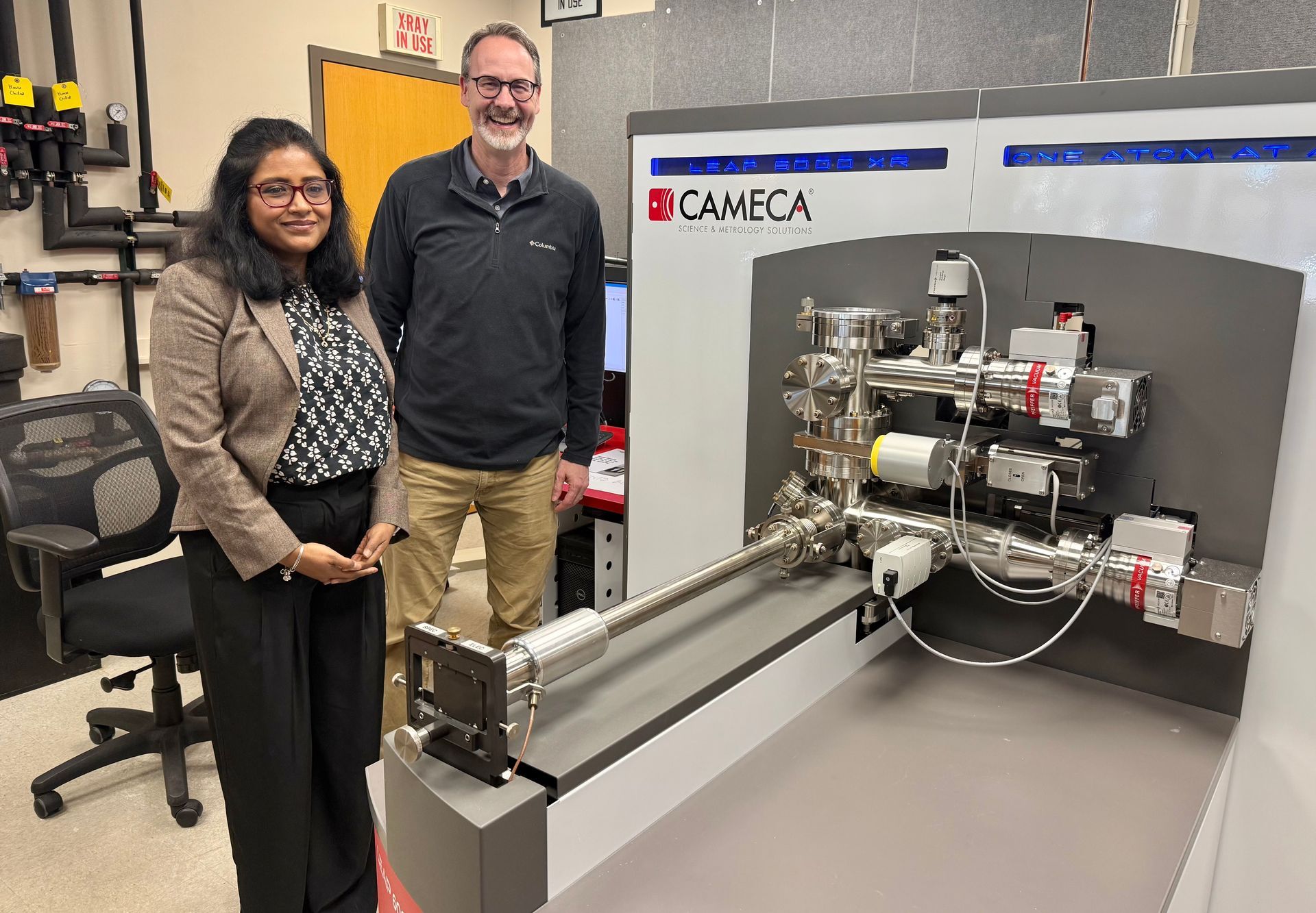 Two people stand beside a CAMECA lab machine in a workshop, with machinery mounted on a gray test bench.
