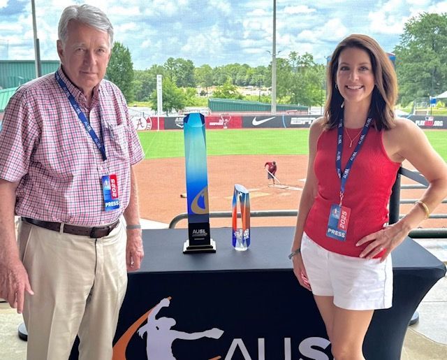 Two people stand by a trophy table at a baseball field, with a stadium and players in the background.
