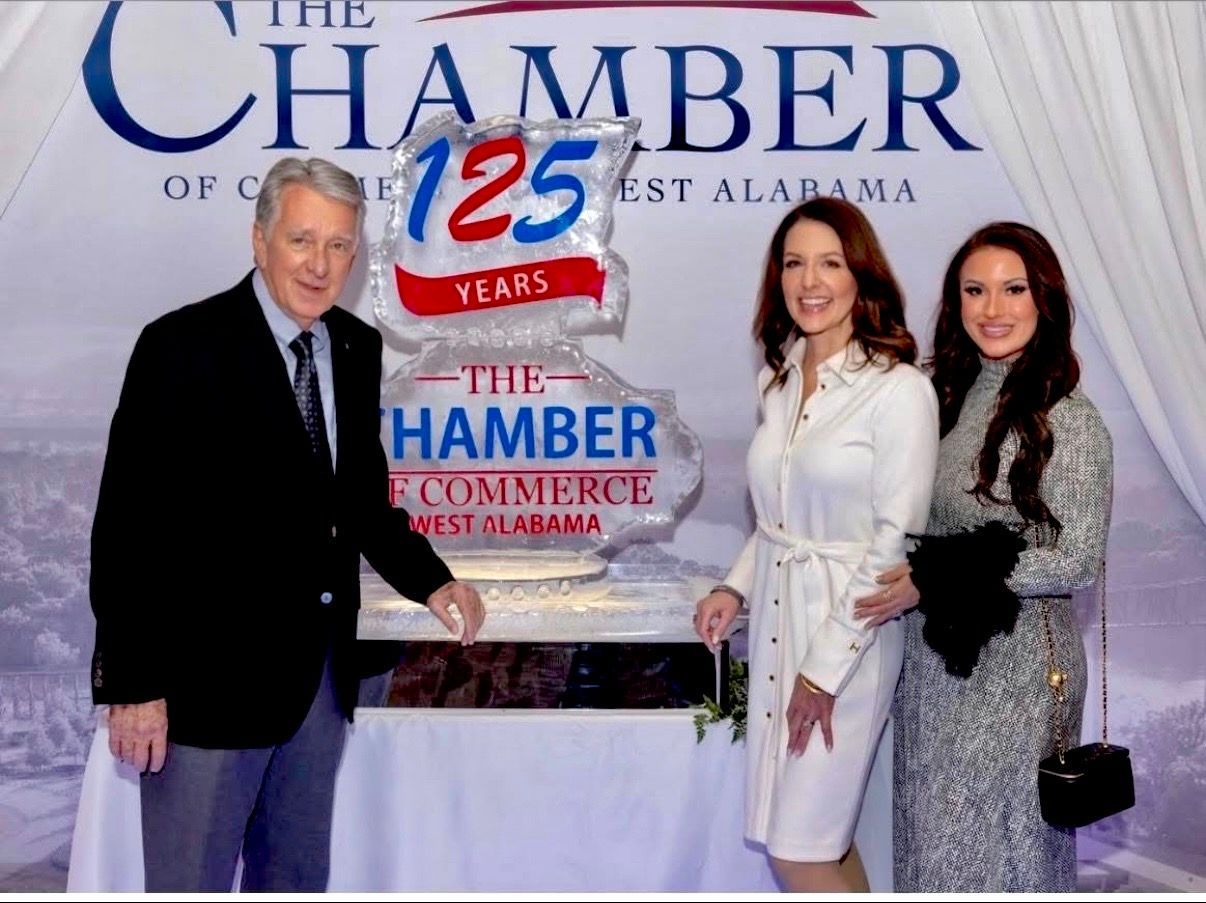 Three people posing beside a Chamber of Commerce 125th anniversary banner and table display