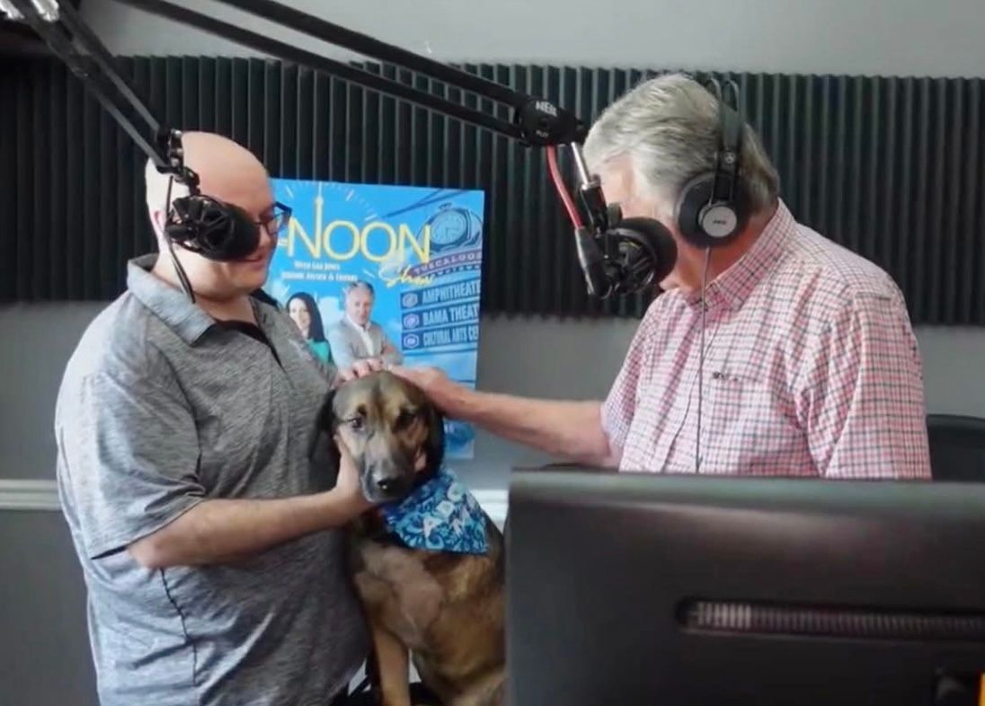 Two hosts in a radio studio pet a small dog between them, with microphones and headphones visible.