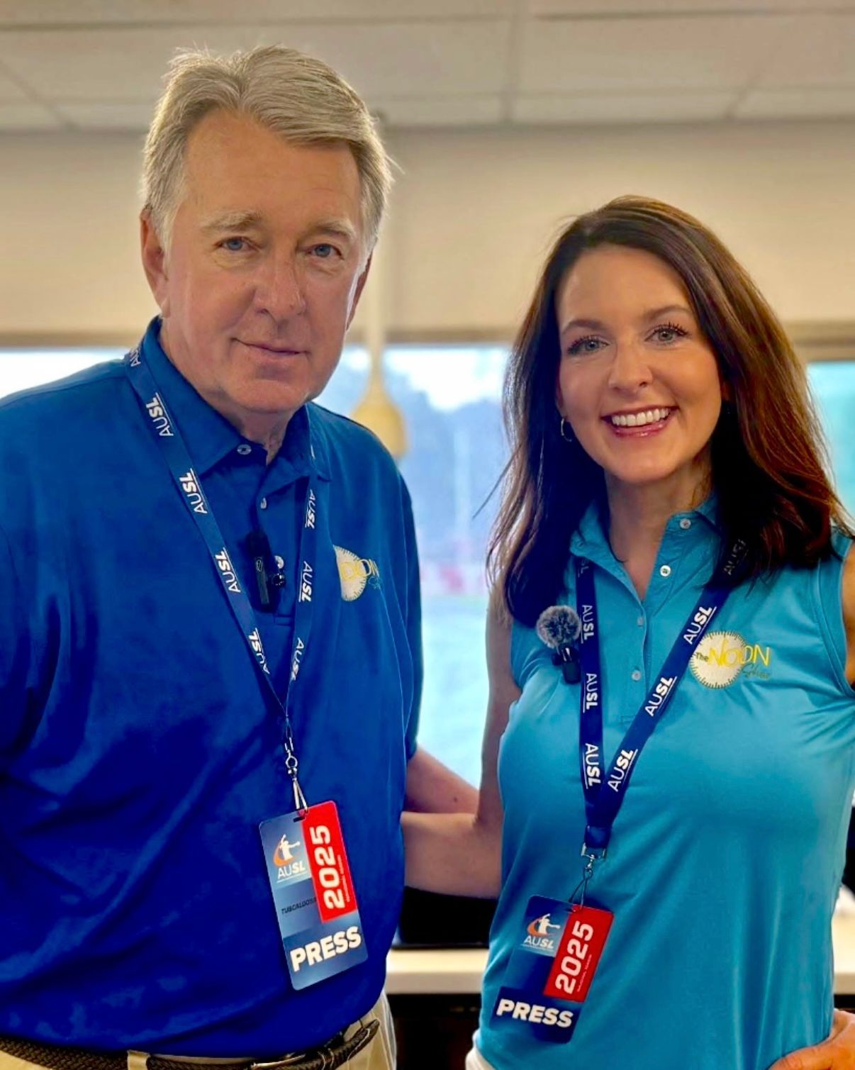 Two people in blue shirts and press lanyards smiling indoors