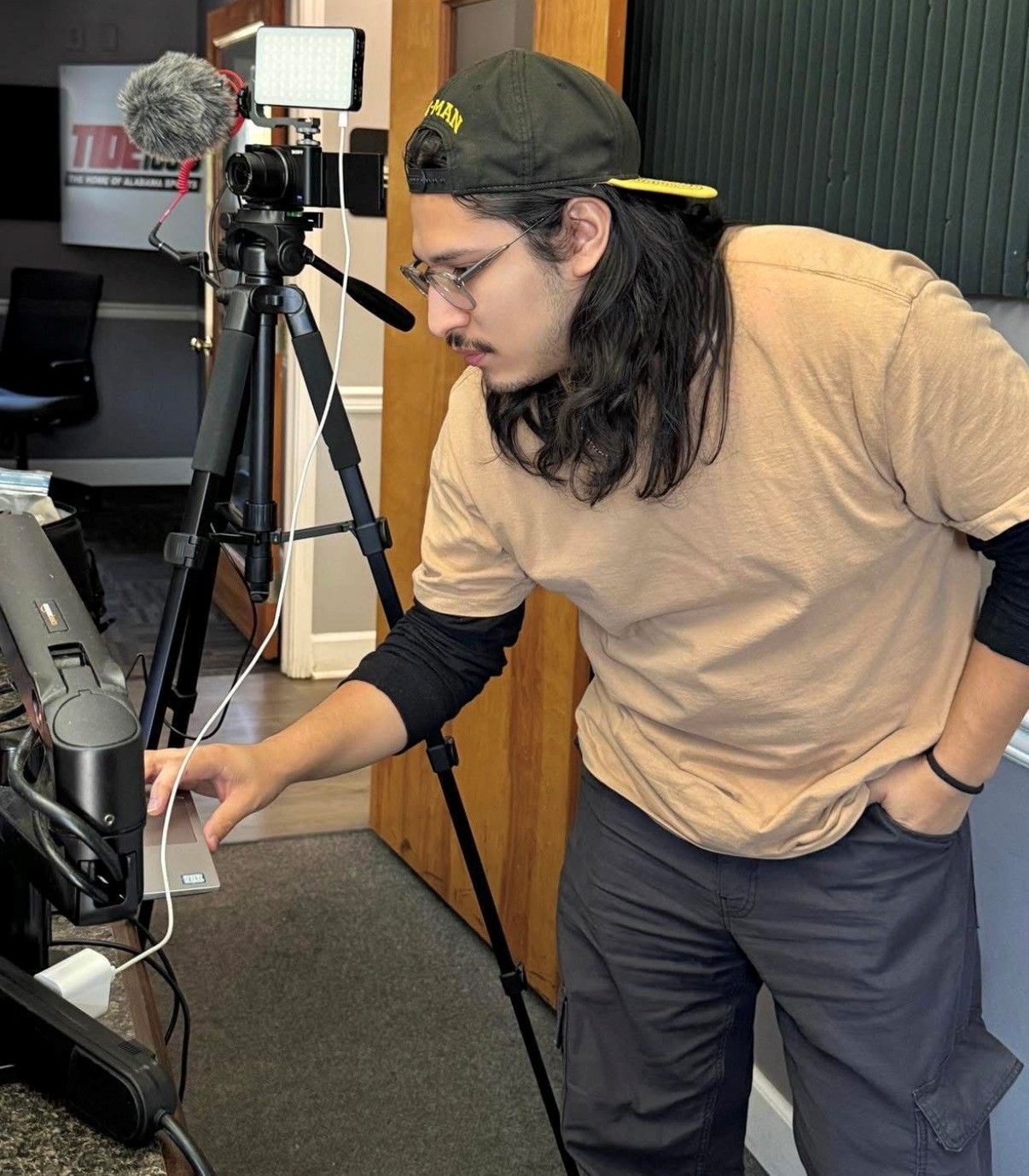 Person adjusting equipment on a tripod in a room with a laptop and camera setup.