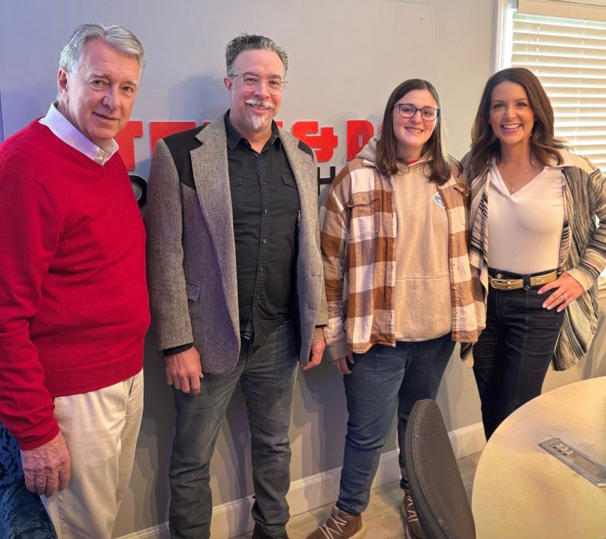 Four people standing together indoors, smiling in front of a wall with a red logo.