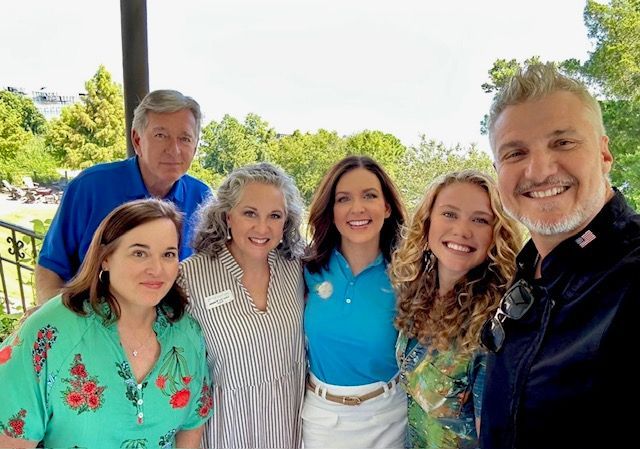 Six smiling people posing outdoors by a railing, with trees and water in the background.
