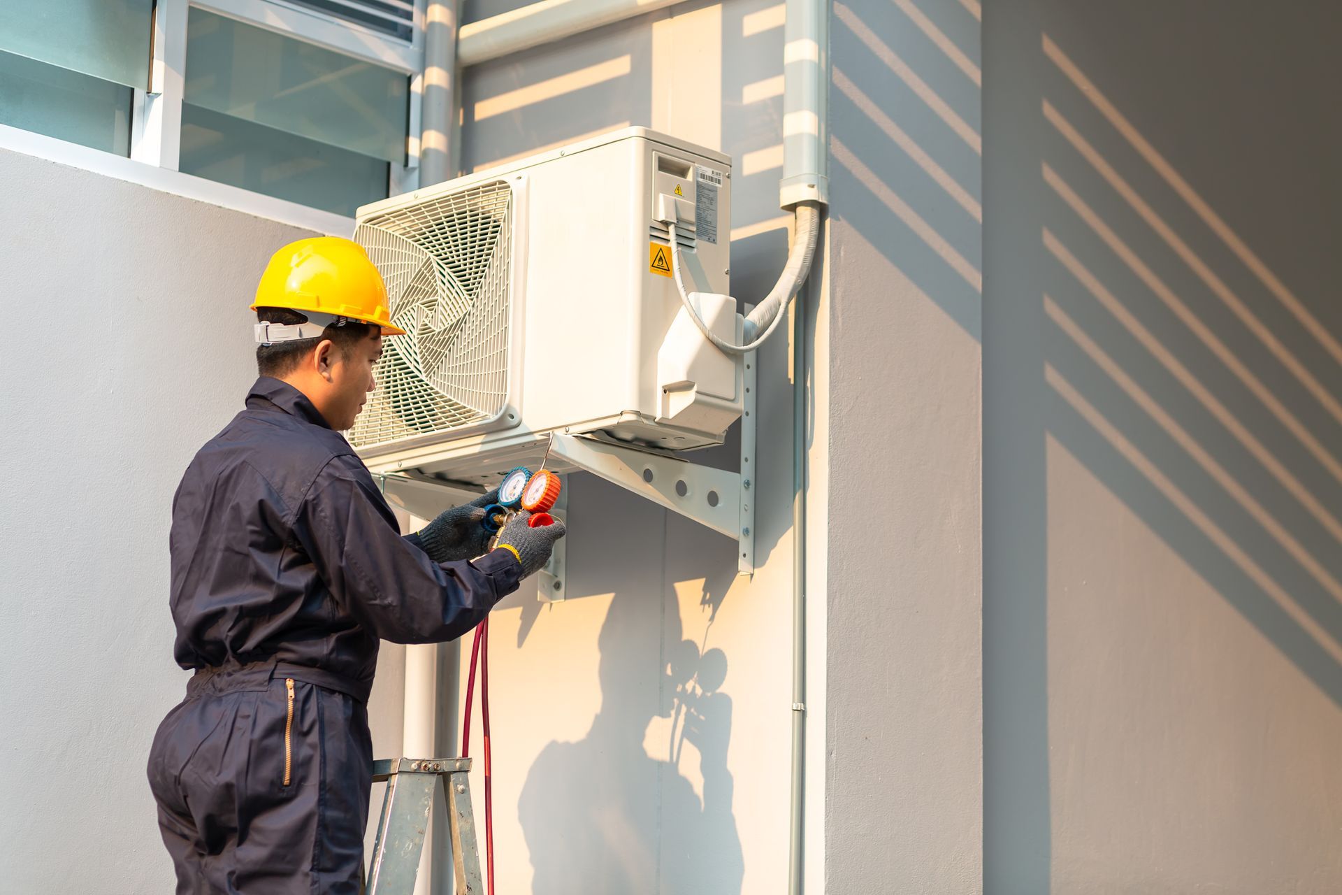HVAC technician in yellow hard hat working on outdoor air conditioner unit on a building.